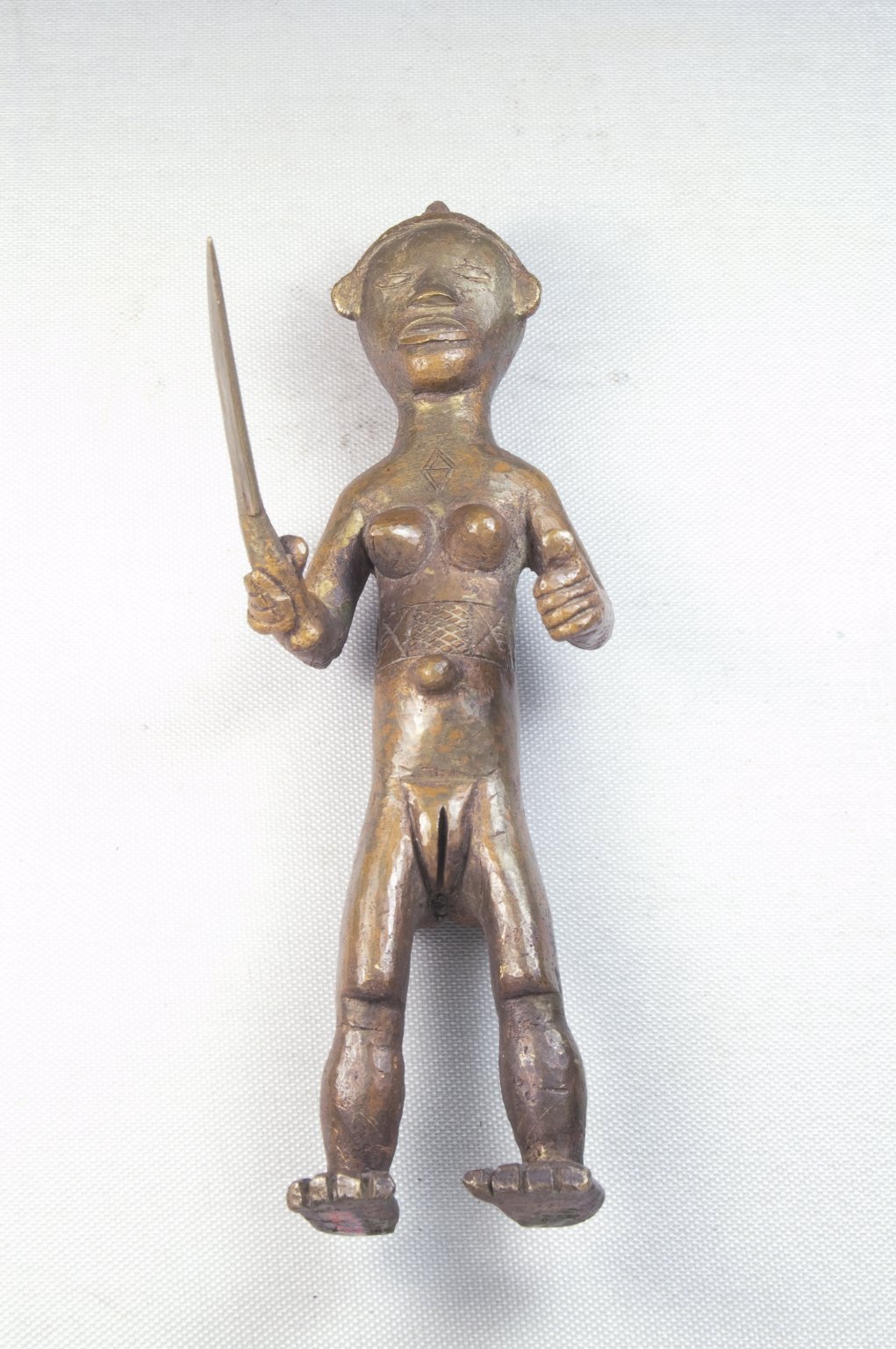 Copper alloy standing female nude holding a large knife. Incised scarification marks on her abdomen and chest. Hair arranged in three ridges: one down center of head; each of other two out from centra