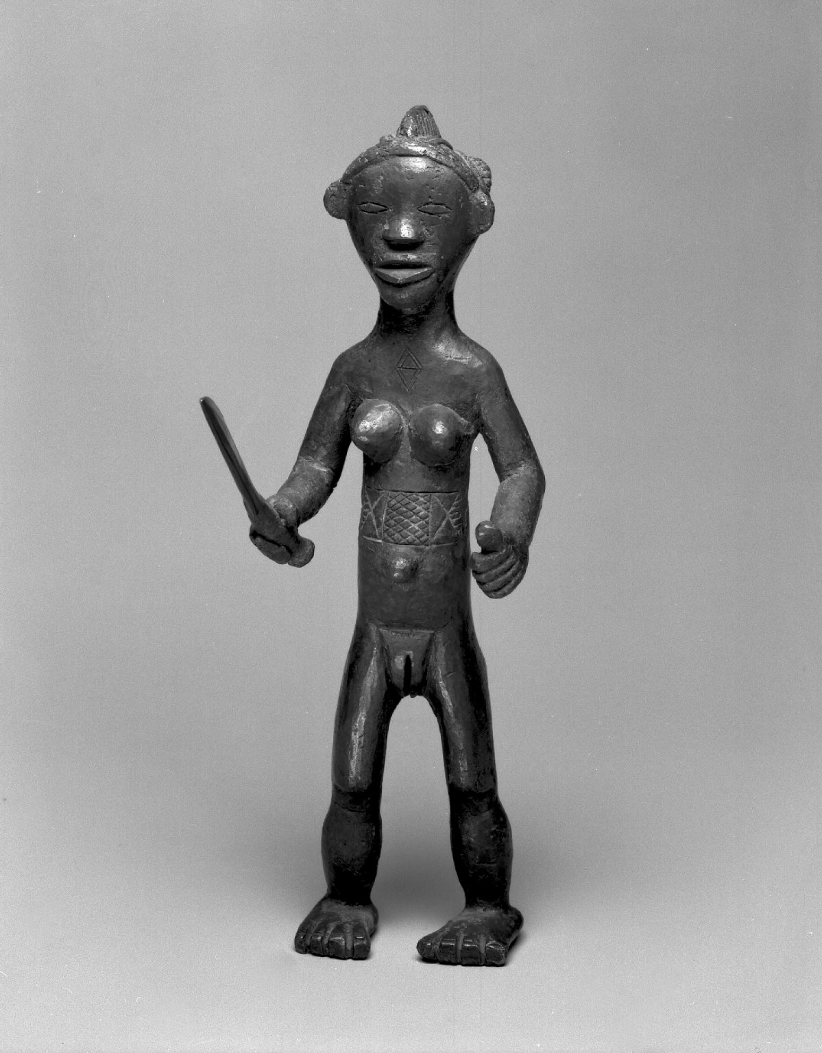 Copper alloy standing female nude holding a large knife. Incised scarification marks on her abdomen and chest. Hair arranged in three ridges: one down center of head; each of other two out from centra