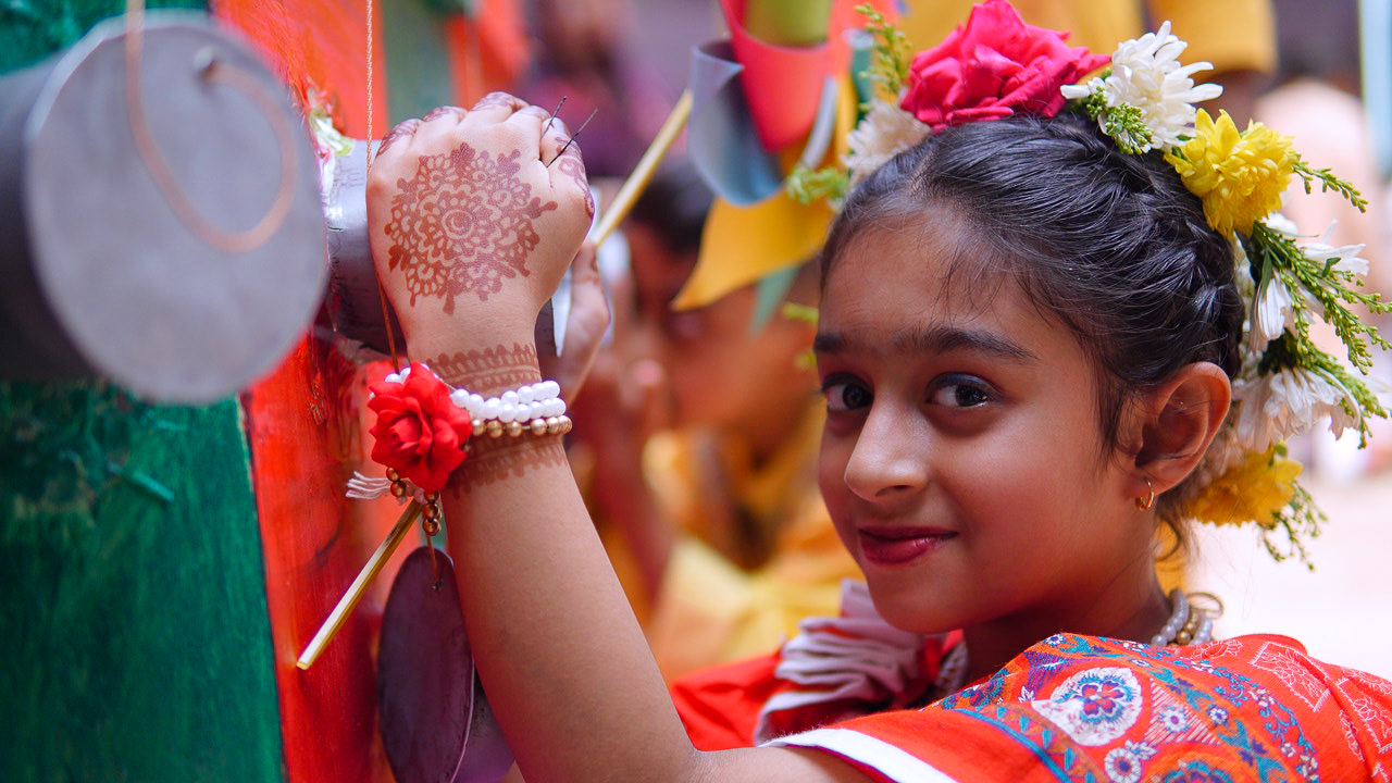 A young girl, adorned in traditional festive attire, looks into the camera with a gentle, knowing smile. Her hands are beautifully decorated with intricate henna (mehndi) patterns, and she wears color