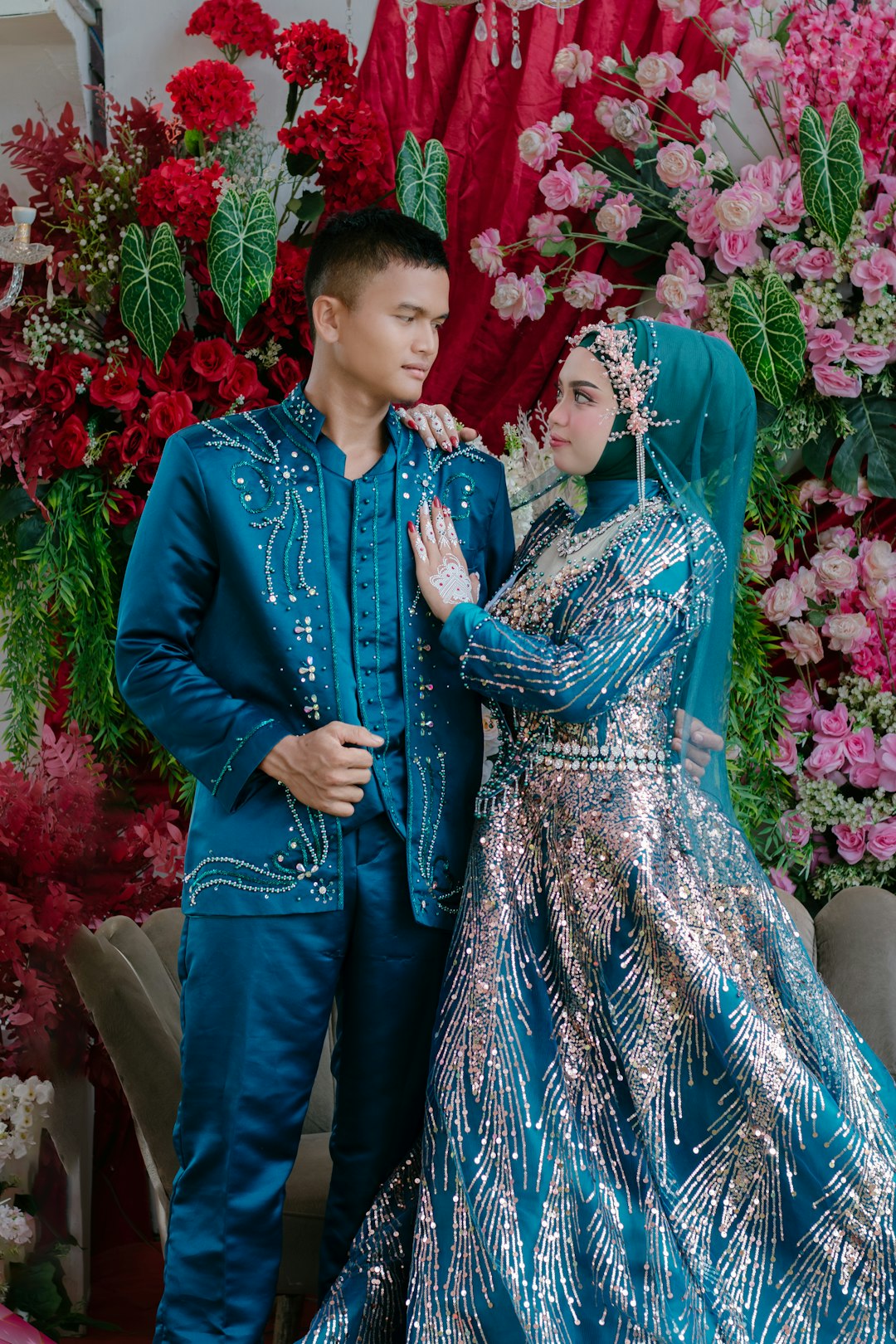 A beautiful and romantic portrait of an Indonesian Muslim couple gazing at each other on their wedding day. The bride looks stunning in a luxurious teal gown heavily adorned with shimmering sequins, complete with a matching hijab and elegant floral h