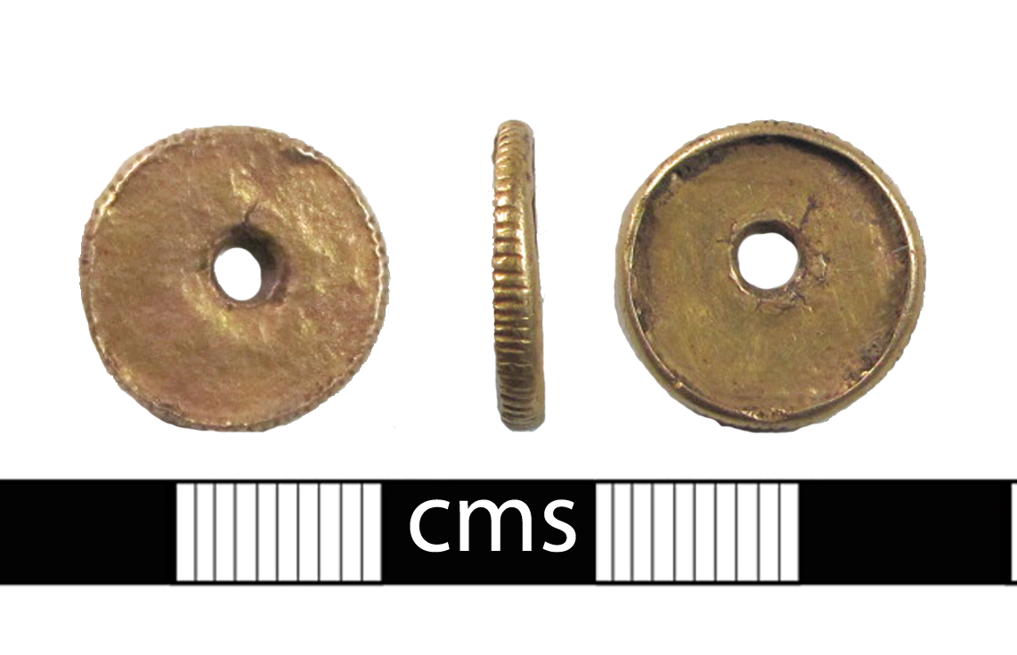A circular disc of gold, with a hand cut 'milled' edge and a piercing through the central flan. The milled edge stands proud of the flan, more so on one side than the other. It is unclear what this it