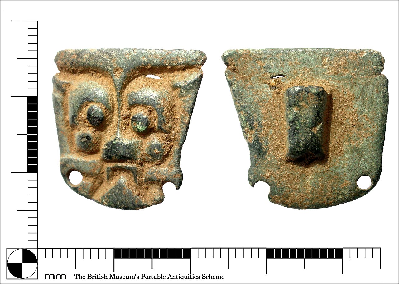 An early incomplete copper alloy mount, possibly a harness mount. It has a humanoid face, large eyebrows and pellet-like eyes, which is repeated in the cheeks! There is a flat nose with a downturned m