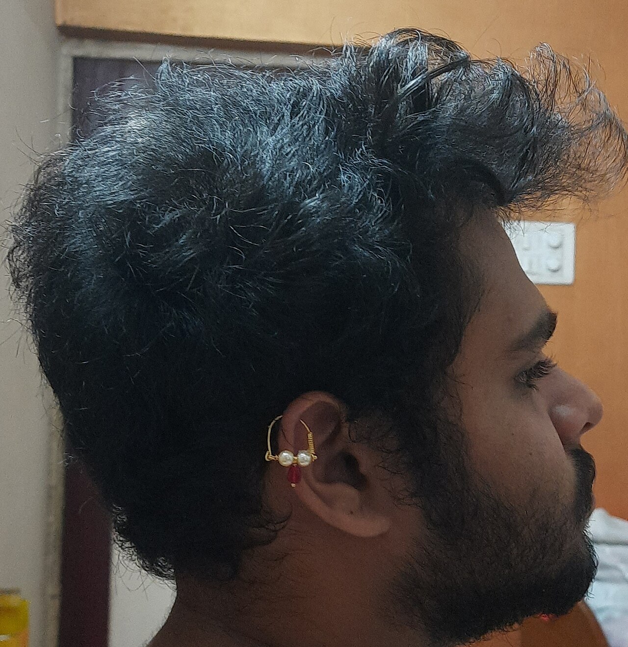 A man wearing a bhikbali on his right ear, the piercing is done in the helix. Two pearls and a red stone can be seen fixed into the bhikbali.