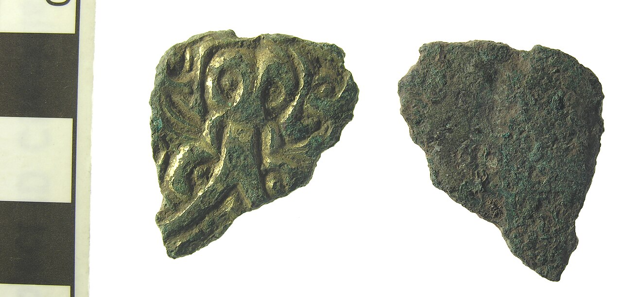 Copper alloy fragment with chip carved decoration and substantial gilding surviving. The decoration comprises a beast or mans head with a bifurcating tongue protruding from the mouth. Two small copper